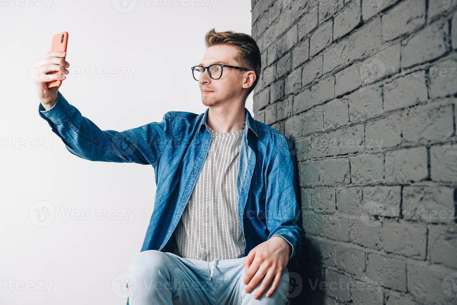 giovane uomo in camicia blu e occhiali che scatta foto selfie sul cellulare su uno sfondo di muro di mattoni neri. copia, spazio vuoto per il testo