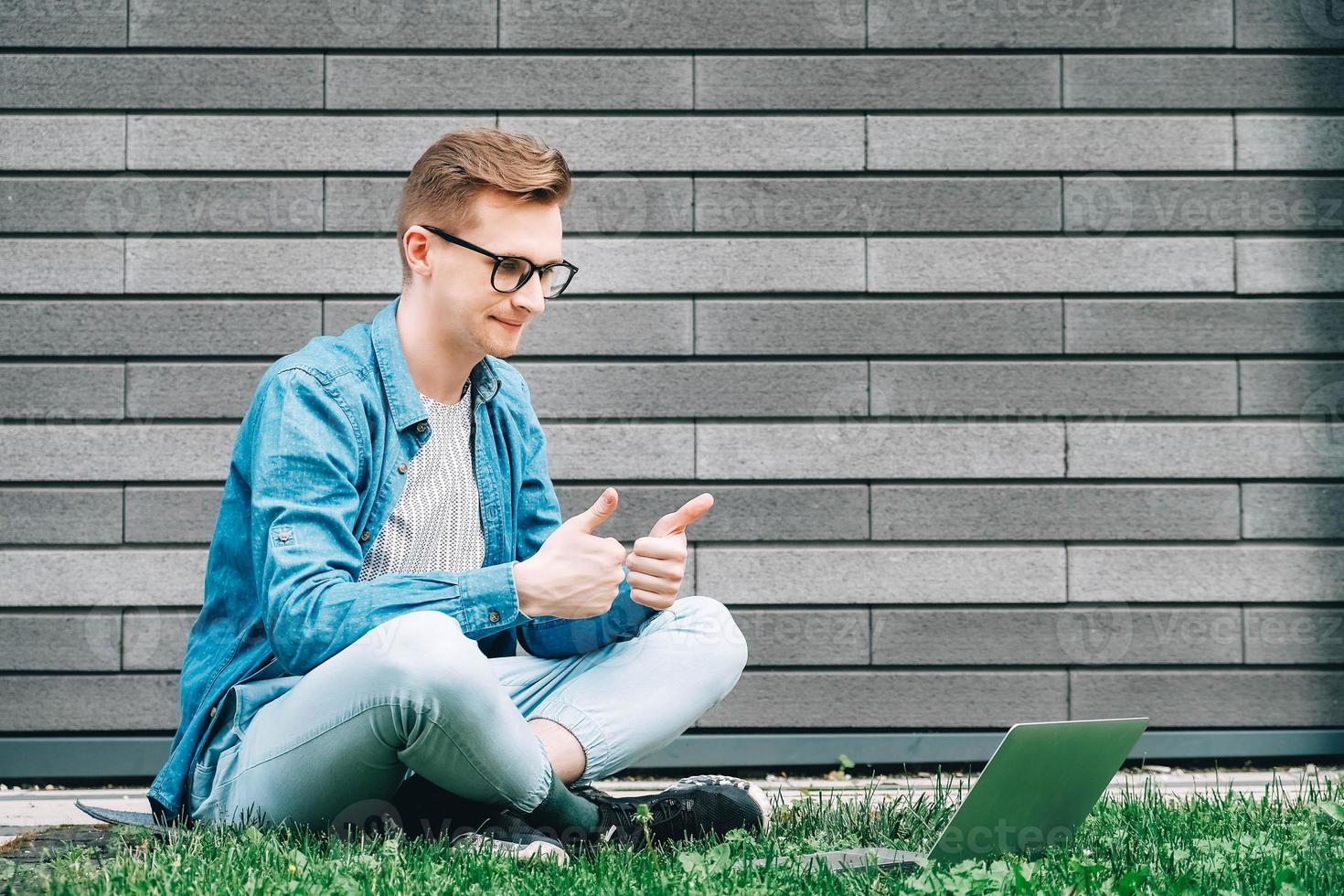 uomo d'affari in camicia, jeans e occhiali seduto sull'erba verde e utilizzando il computer portatile sullo sfondo di un muro grigio di casa foto