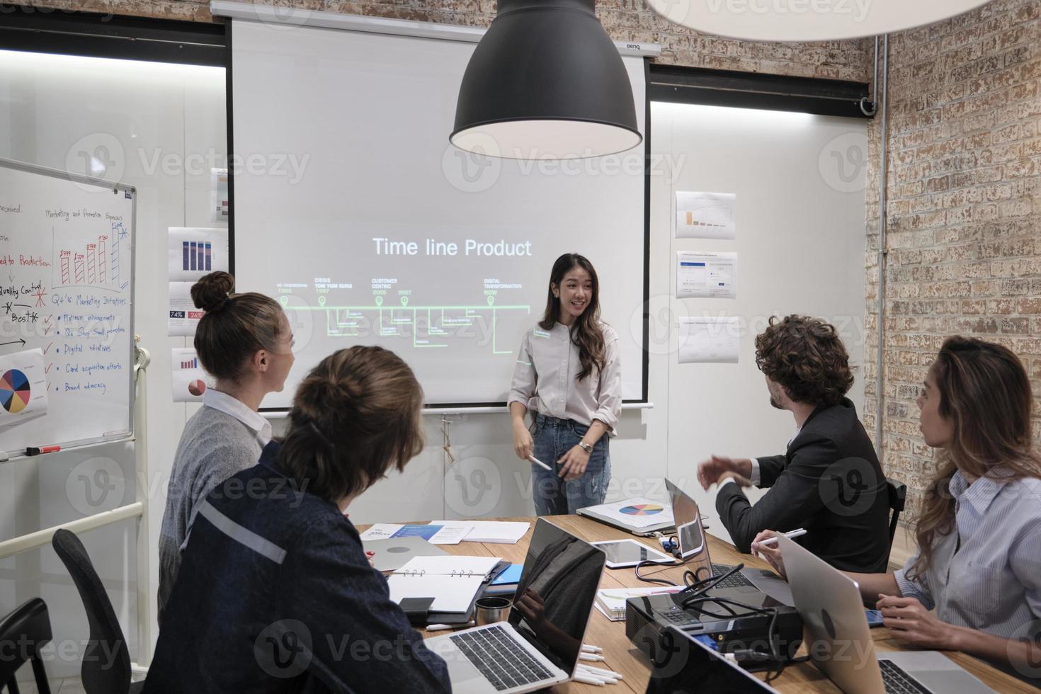 un simpatico personale femminile asiatico in piedi e presentando la strategia di marketing aziendale al suo capo e ai colleghi multirazziali, parlando fiducioso nella sala conferenze della riunione di brainstorming presso l'ufficio aziendale. foto