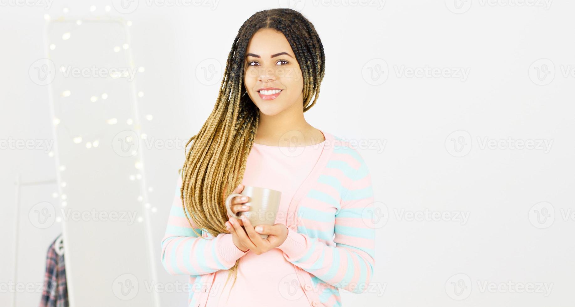 giovane donna nera con acconciatura afro sorridente. ragazza con tazza di bevanda calda che indossa un abito rosa. girato in studio. foto