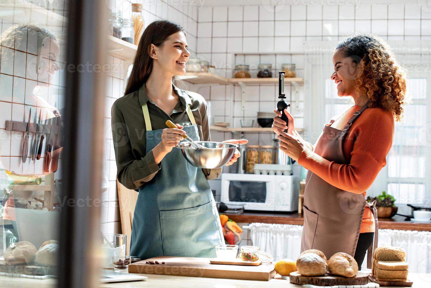 felici due amiche interrazziali che trasmettono video in diretta con il telefono cellulare da condividere online mentre cucinano in cucina a casa foto