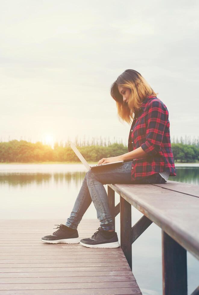 giovane donna felice hipster che lavora con il suo computer portatile seduto sul molo, rilassante godere con la natura. foto