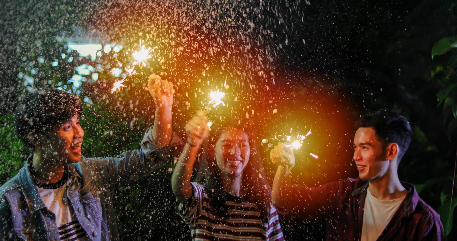 gruppo asiatico di amici che hanno barbecue in giardino all'aperto ridendo con bevande alcoliche di birra e mostrando un gruppo di amici che si divertono con le stelle filanti la notte foto