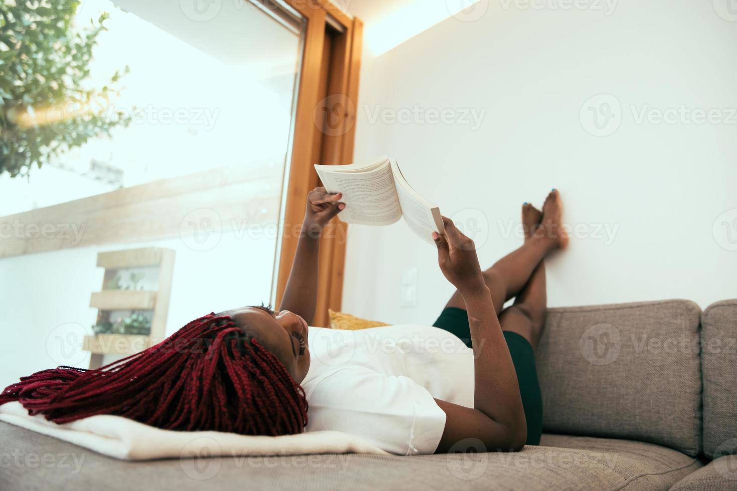 donna nera sdraiata sul divano a leggere un libro con i piedi sul muro foto