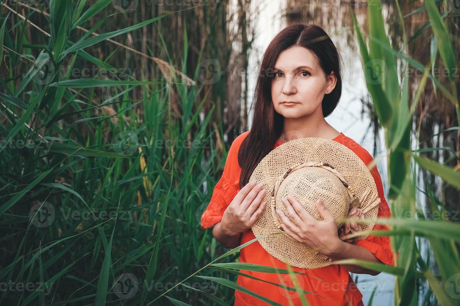 ritratto di una bella donna con un cappello di paglia vestito con abiti rossi su uno sfondo di canne e lago foto