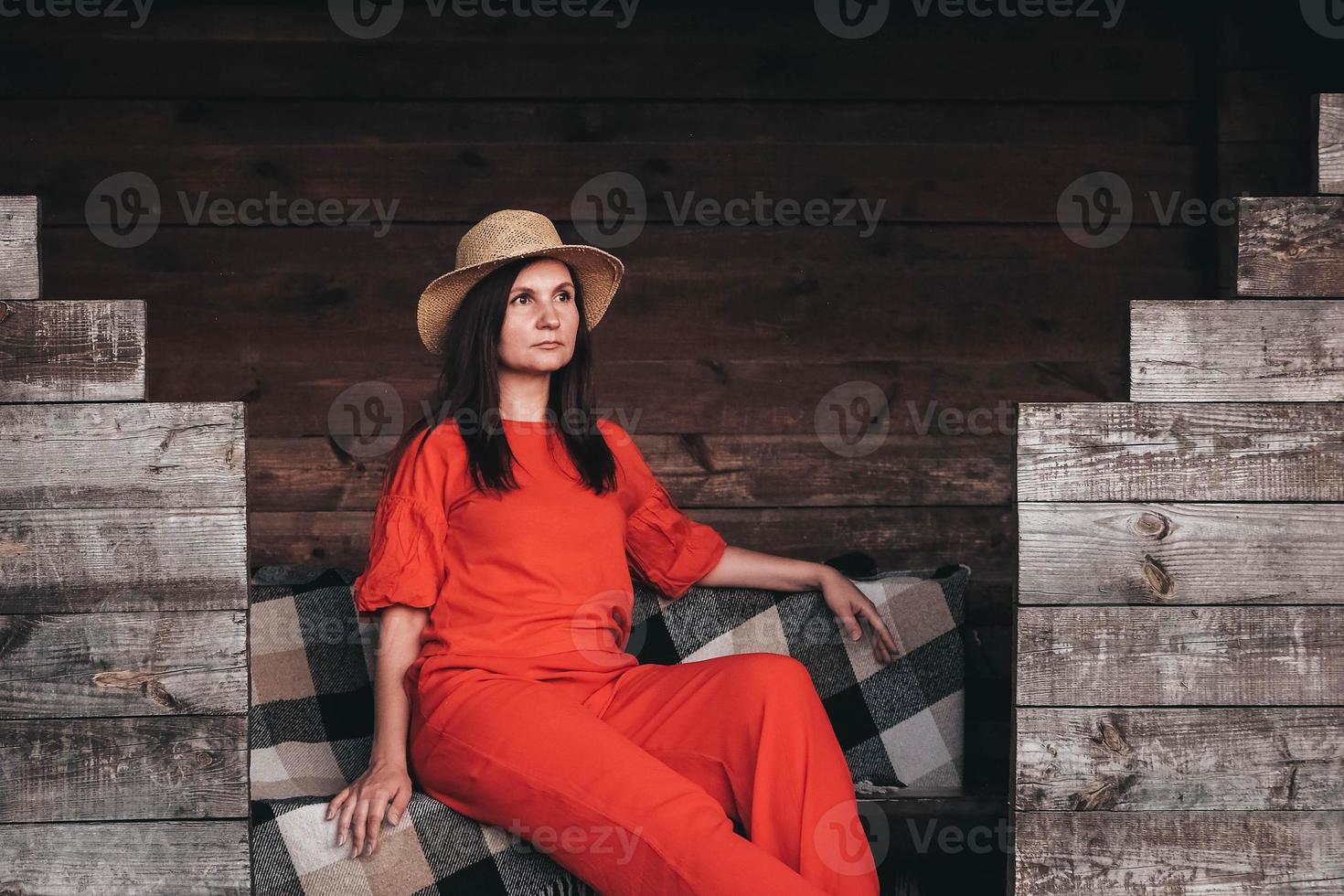 bella donna con un cappello di paglia vestita con abiti rossi seduta sullo sfondo di una casa di legno foto