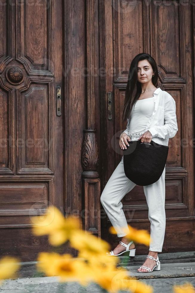 donna che indossa abiti bianchi, tenendo in mano una borsa a maglia sullo sfondo di vecchie porte di legno. copia, spazio vuoto per il testo foto