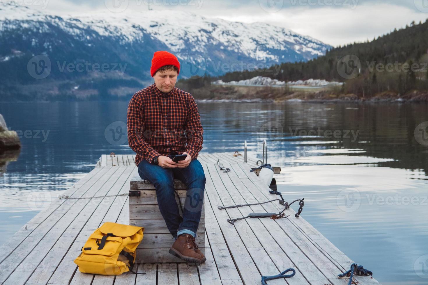 uomo che guarda nello smartphone con uno zaino giallo che indossa un cappello rosso seduto sul molo di legno sullo sfondo della montagna e del lago foto
