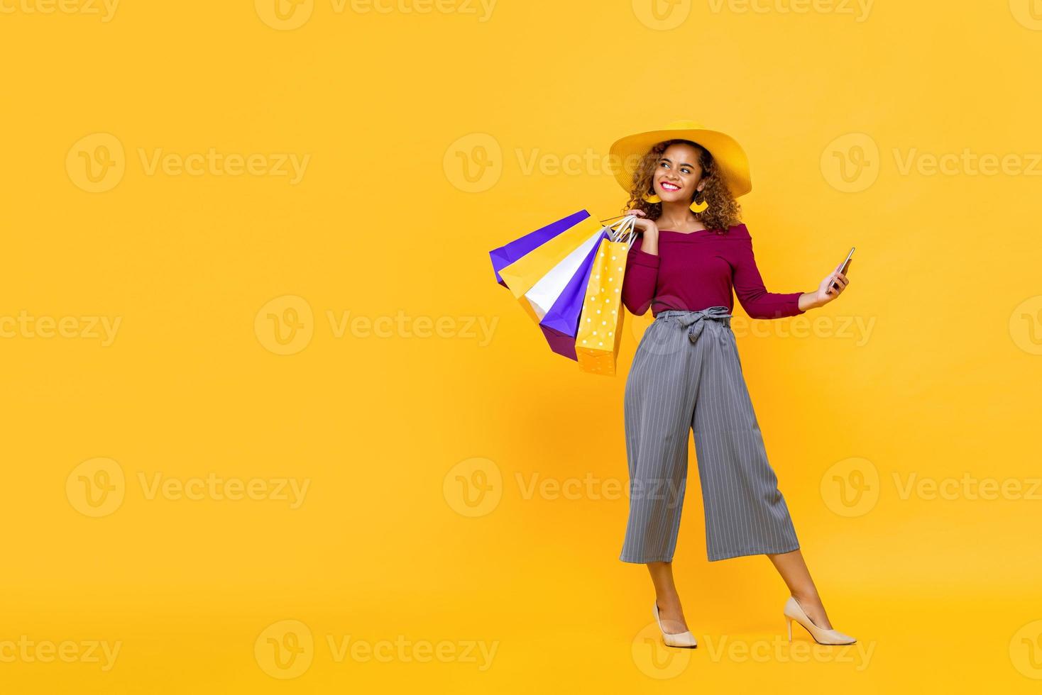 ritratto a figura intera di donna afroamericana sorridente che tiene borse della spesa e telefono cellulare mentre guarda lateralmente per copiare lo spazio su sfondo giallo foto