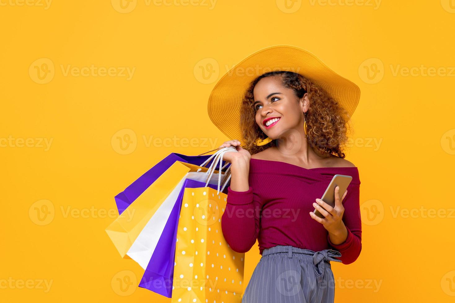 donna di razza mista sorridente alla moda con borse della spesa colorate e telefono cellulare isolato su sfondo giallo per conept vendita estiva foto