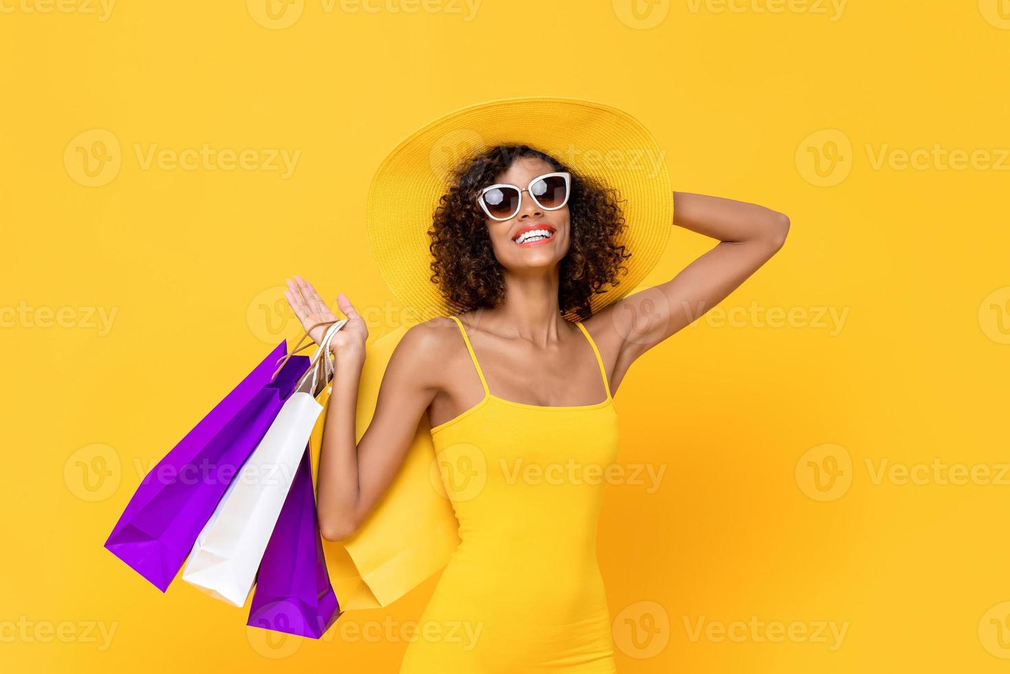 ritratto estivo di allegra sorridente donna afroamericana che indossa occhiali da sole tenendo il cappello e borse della spesa in studio isolato sfondo giallo foto