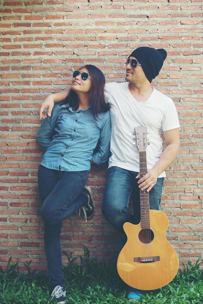 la coppia di hipster si abbraccia, distoglie lo sguardo e sorride mentre è in piedi all'aperto a suonare la chitarra contro un muro di mattoni, frequentando trascorsi molto tempo insieme. foto