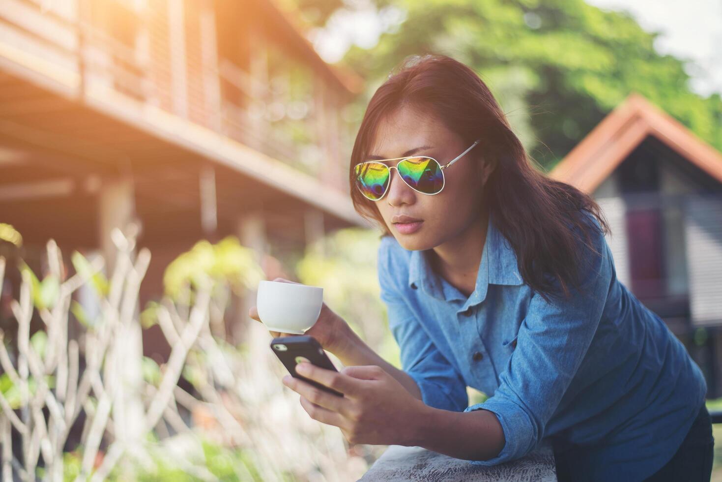 giovane donna hipster che guarda il suo telefono mentre tiene una tazza di caffè al mattino e sorride. concetto di vacanza rilassante. foto