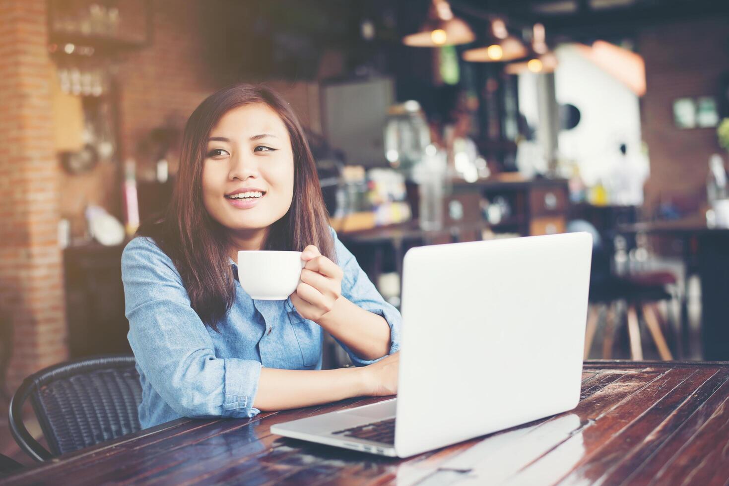 bella giovane donna hipster seduta in una caffetteria, con in mano una tazza di caffè, rilassarsi e giocare con il suo computer portatile, distogliendo lo sguardo, felice e divertente. concetto di stile di vita. foto