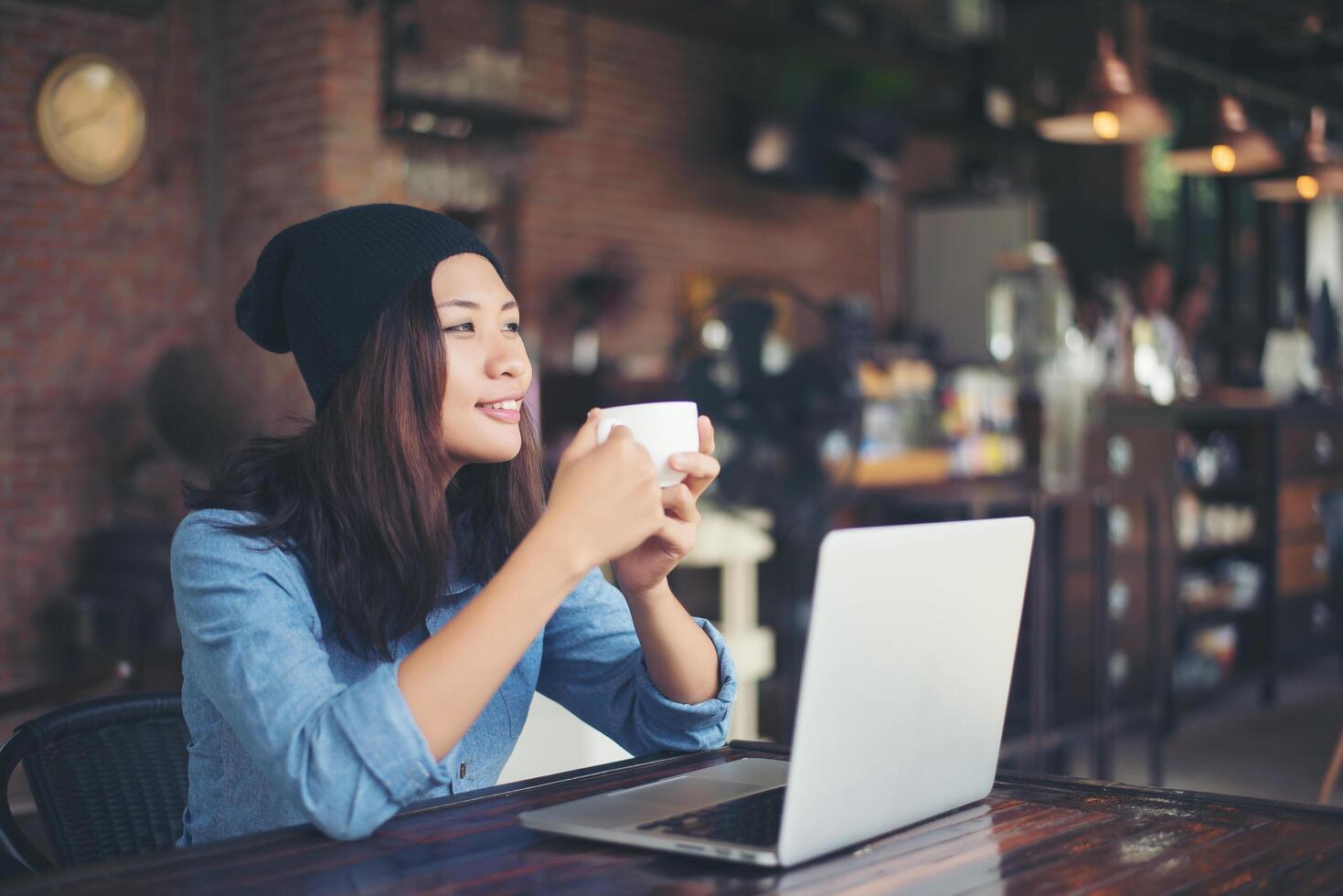 bella giovane donna hipster seduta in una caffetteria, con in mano una tazza di caffè, rilassarsi e giocare con il suo computer portatile, distogliendo lo sguardo, felice e divertente. concetto di stile di vita. foto