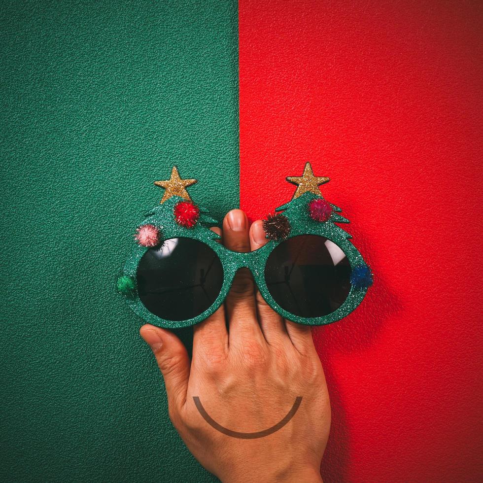 occhiali di natale che decorazione con albero di natale e palla rossa a portata di mano su sfondo verde e rosso foto