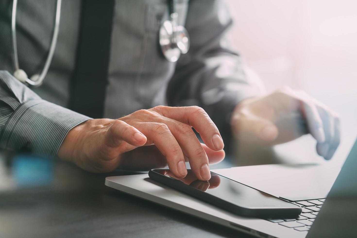 primo piano di un medico intelligente che lavora con il telefono cellulare e il computer portatile e lo stetoscopio sulla scrivania di legno scuro foto