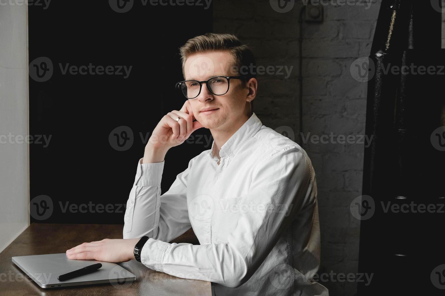 Ritratto di giovane uomo che indossa una camicia bianca e occhiali da vista seduto con il laptop e lavorando nella caffetteria. concetto di libero professionista. copia, spazio vuoto per il testo foto