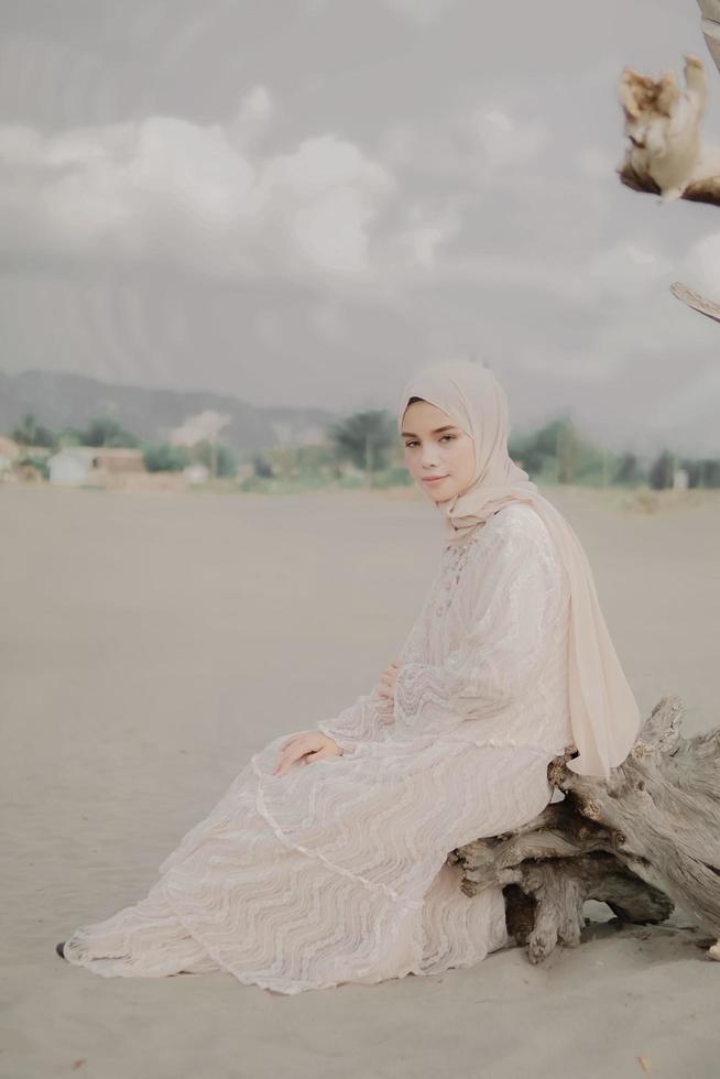 bella modella islamica che indossa la moda hijab, un moderno abito da sposa per donna musulmana seduta sulla sabbia e sulla spiaggia. ritrarre un modello di ragazza asiatica usando l'hijab divertendosi in spiaggia con alberi foto