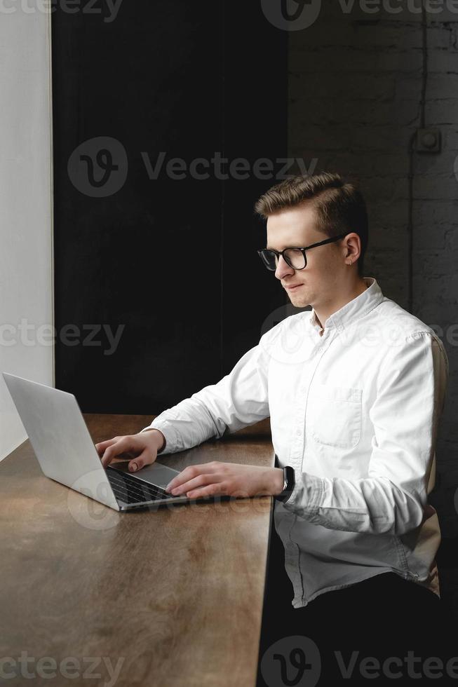 giovane uomo che indossa una camicia bianca e occhiali da vista seduto con il laptop e lavora nella caffetteria foto