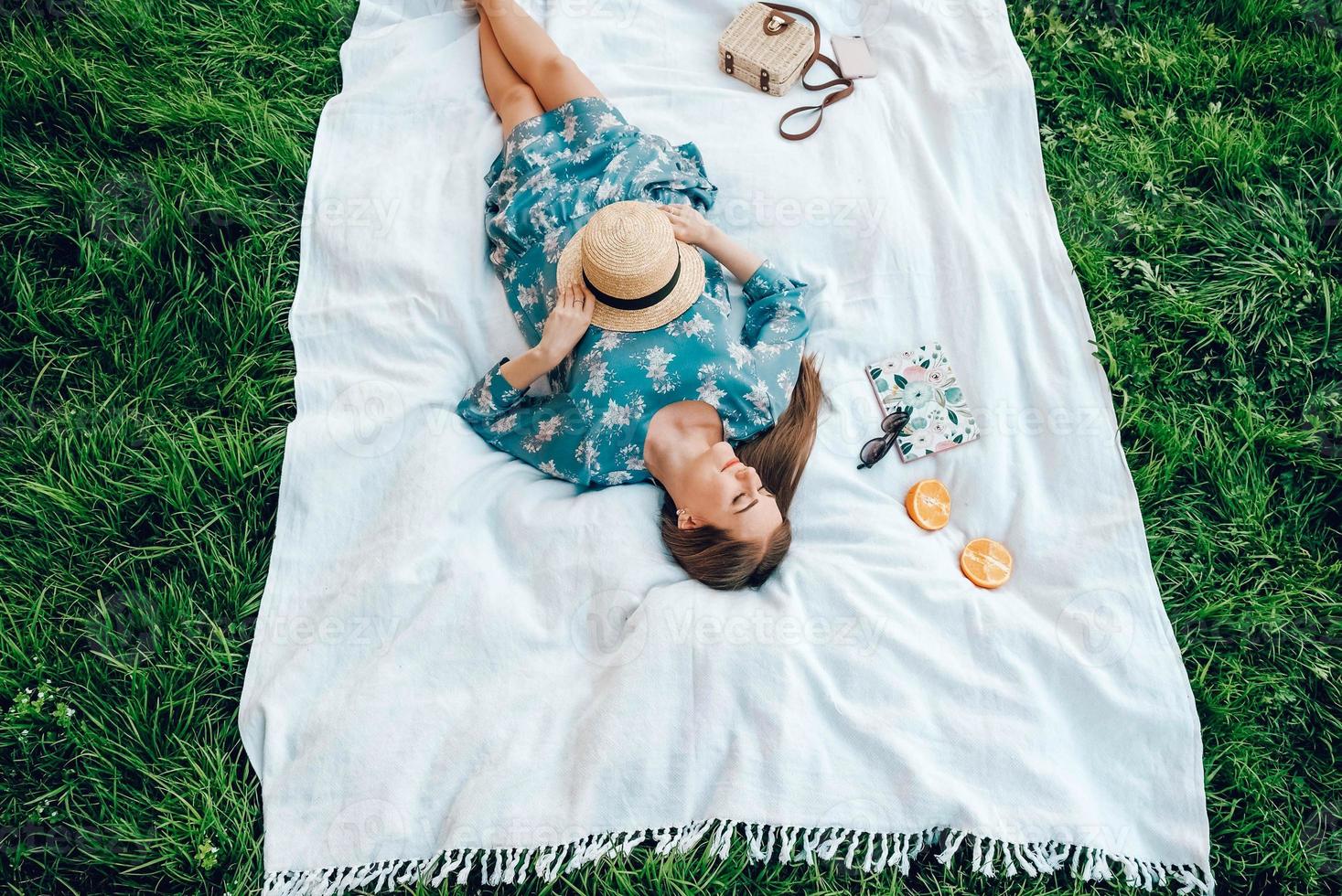 donna con un vestito colorato e un cappello di paglia giace su una coperta bianca su uno sfondo di erba verde, accessori da donna, ora legale foto