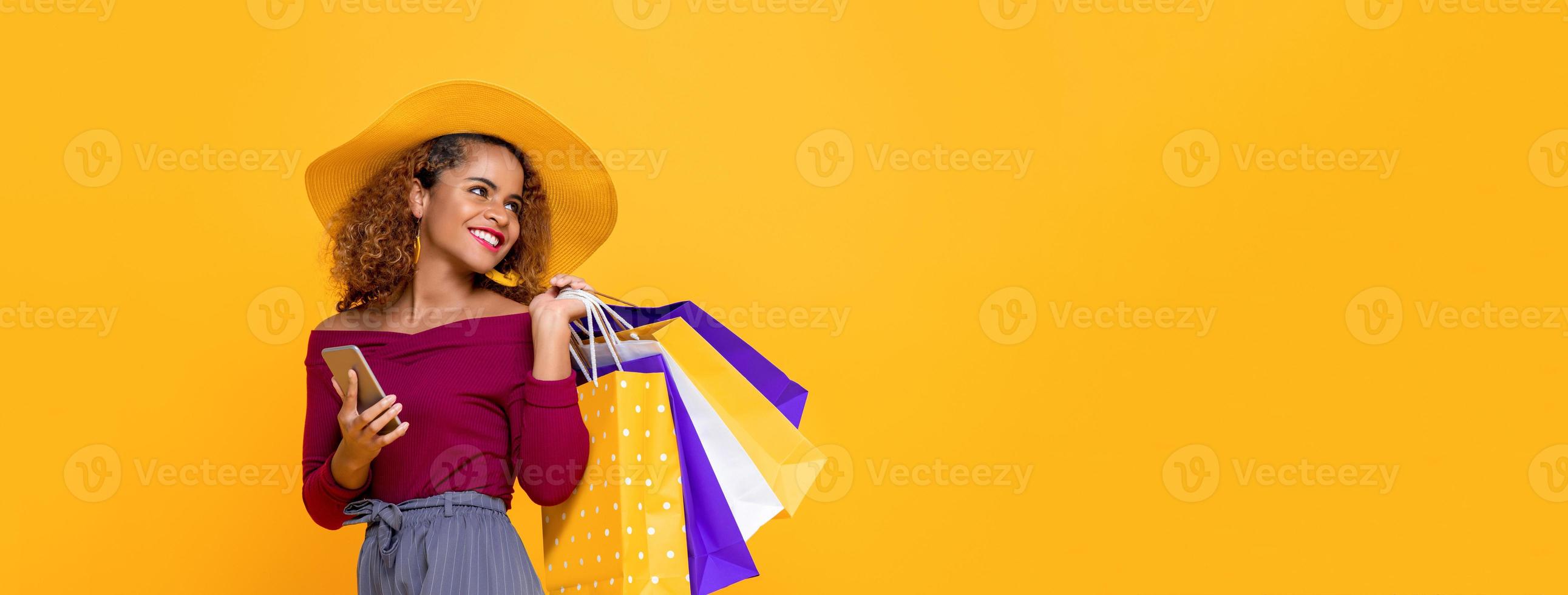 donna di razza mista sorridente alla moda con borse della spesa colorate e telefono cellulare isolato su sfondo giallo per conept vendita estiva foto