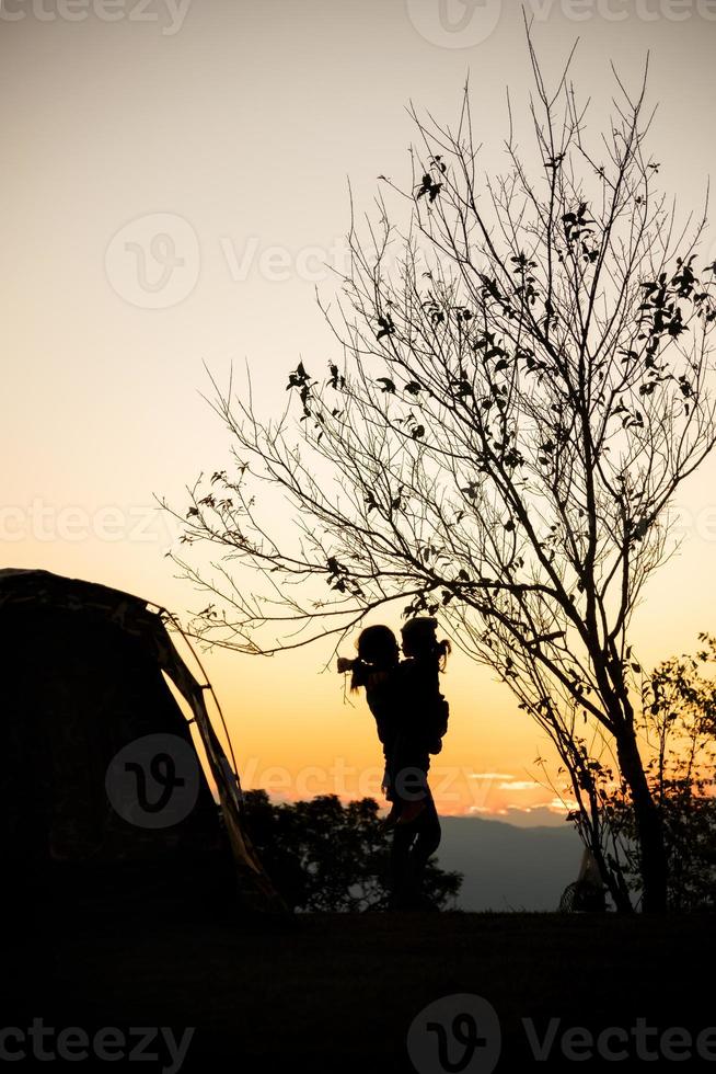 sagoma di madre con sua figlia vicino alla tenda e albero al tramonto foto