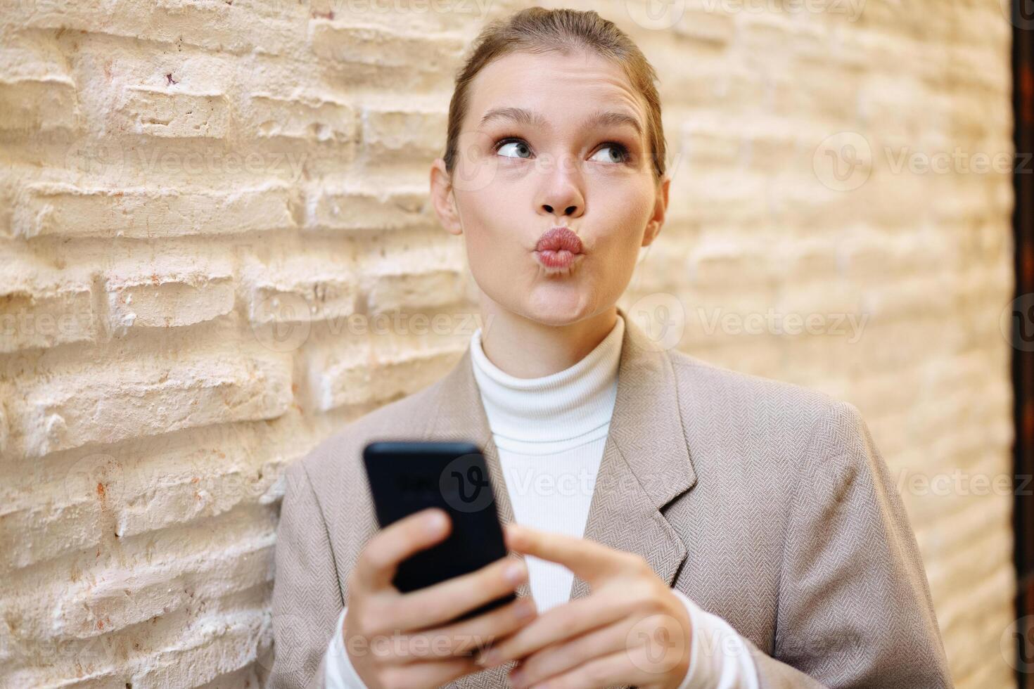 un' giovane donna con gioia e scherzosamente in posa con sua Telefono contro un' strutturato parete foto