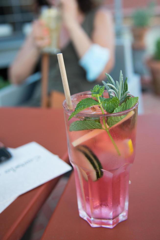 acqua di cetriolo con frutti di bosco e fetta di limone, dal colore rosa e una cannuccia di cartone. donna sullo sfondo con un altro bicchiere. foto