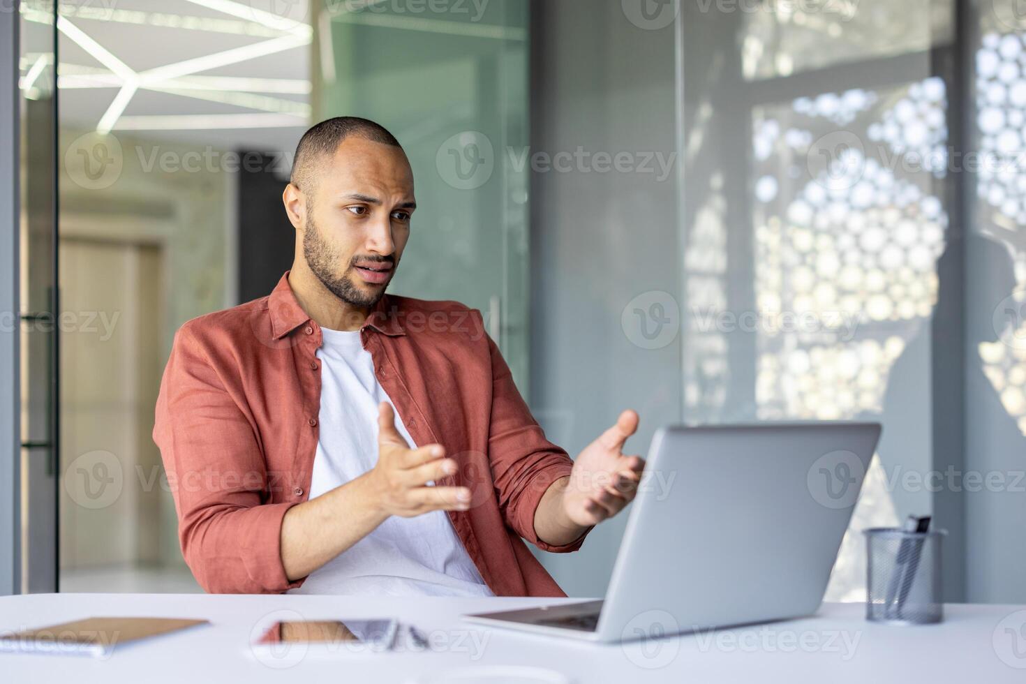 giovane professionale utilizzando il computer portatile Esprimere a gesti espressivamente durante in linea incontro. confuso e frustrato sembra, mostrando preoccupazione. moderno ufficio ambientazione indica tecnologia e comunicazione sfide. foto