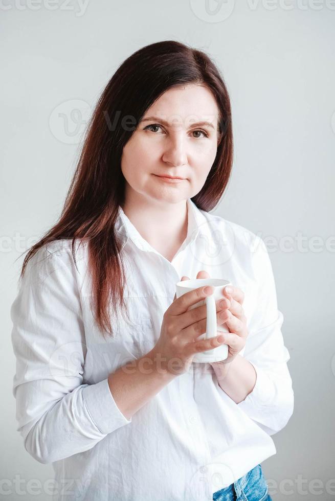 bella donna in una camicia bianca che tiene una tazza di ceramica bianca su sfondo bianco. donna acqua potabile caffè tè dalla tazza bianca. copia, spazio vuoto per il testo foto