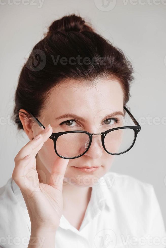 ritratto di una bella donna seria con occhiali ottici e una camicia bianca guardando la telecamera su uno sfondo bianco foto