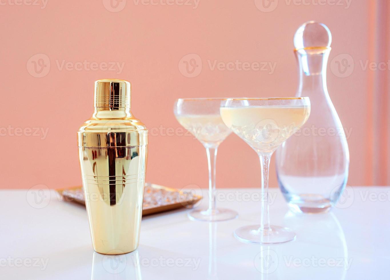 shaker dorato, decanter in vetro, bicchieri eleganti con cocktail e ghiaccio su fondo beige. alcol, festa, hotel, concetto di bar foto