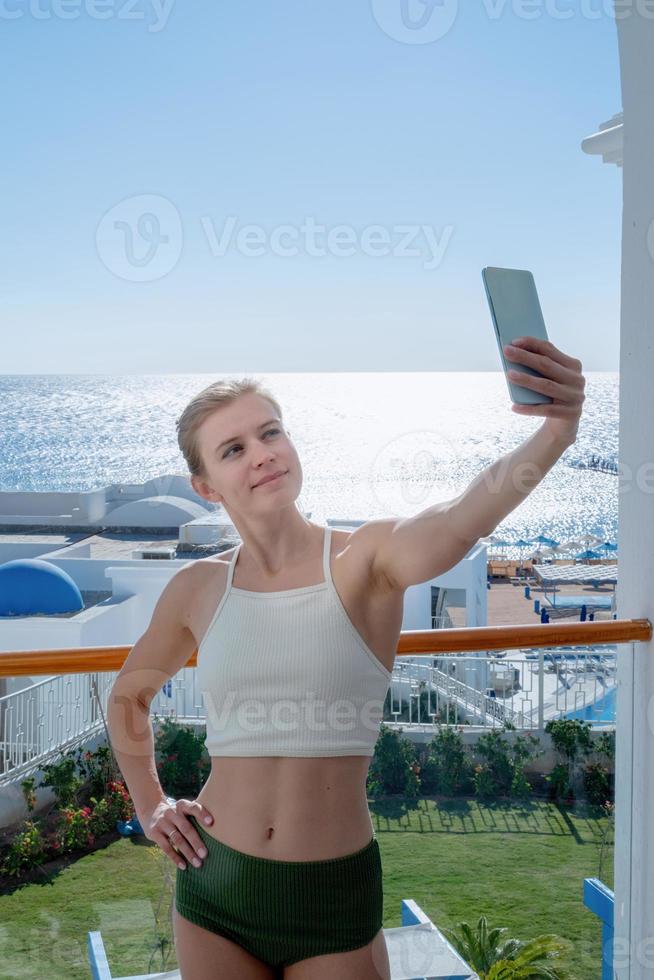 donna felice in costume da bagno che si fa selfie in piedi sul balcone della camera d'albergo foto