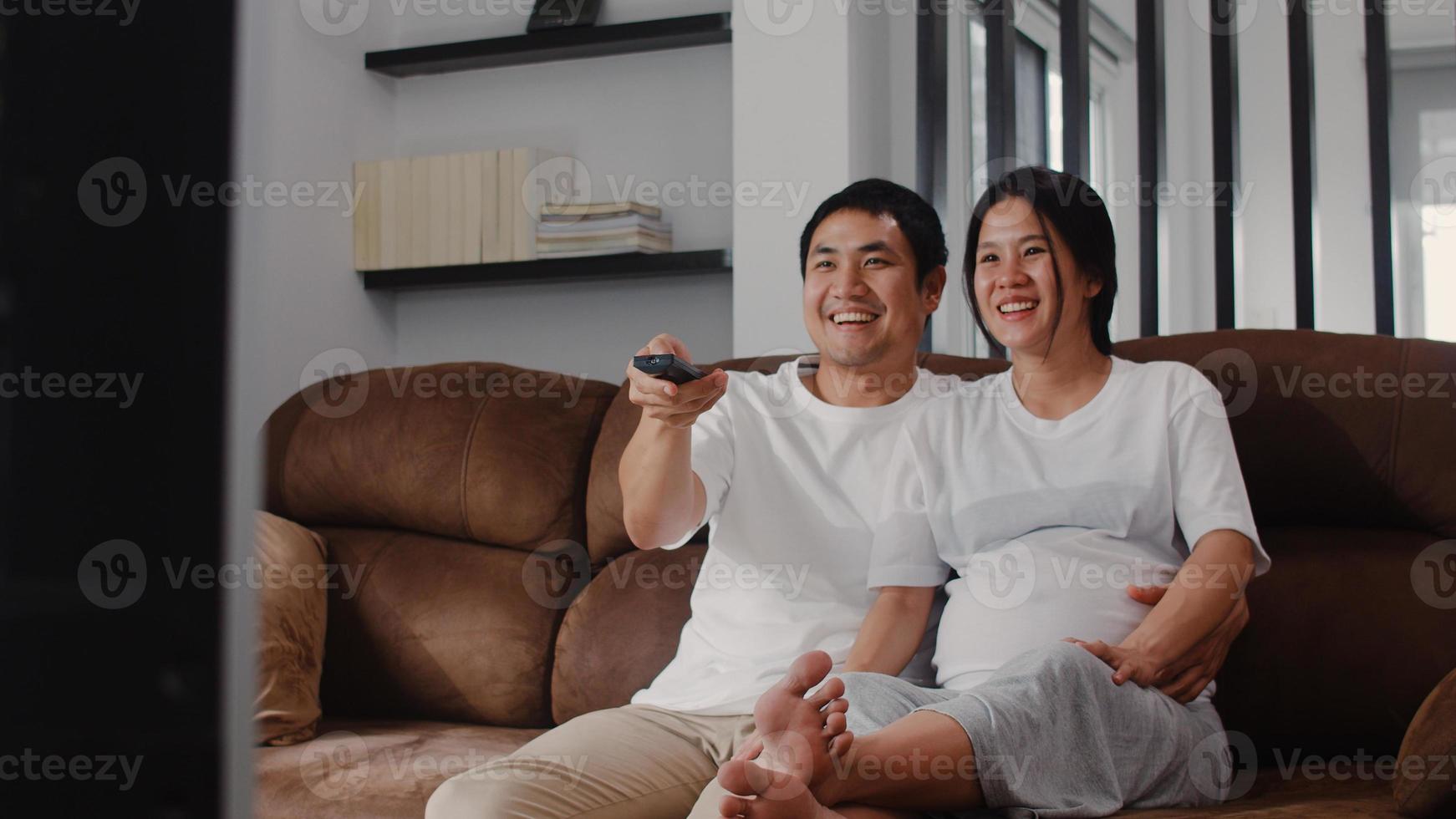 giovane coppia asiatica incinta che guarda la tv. mamma e papà si sentono felici sorridenti pacifici mentre si prendono cura del bambino, gravidanza sdraiato sul divano nel soggiorno a casa concetto. foto