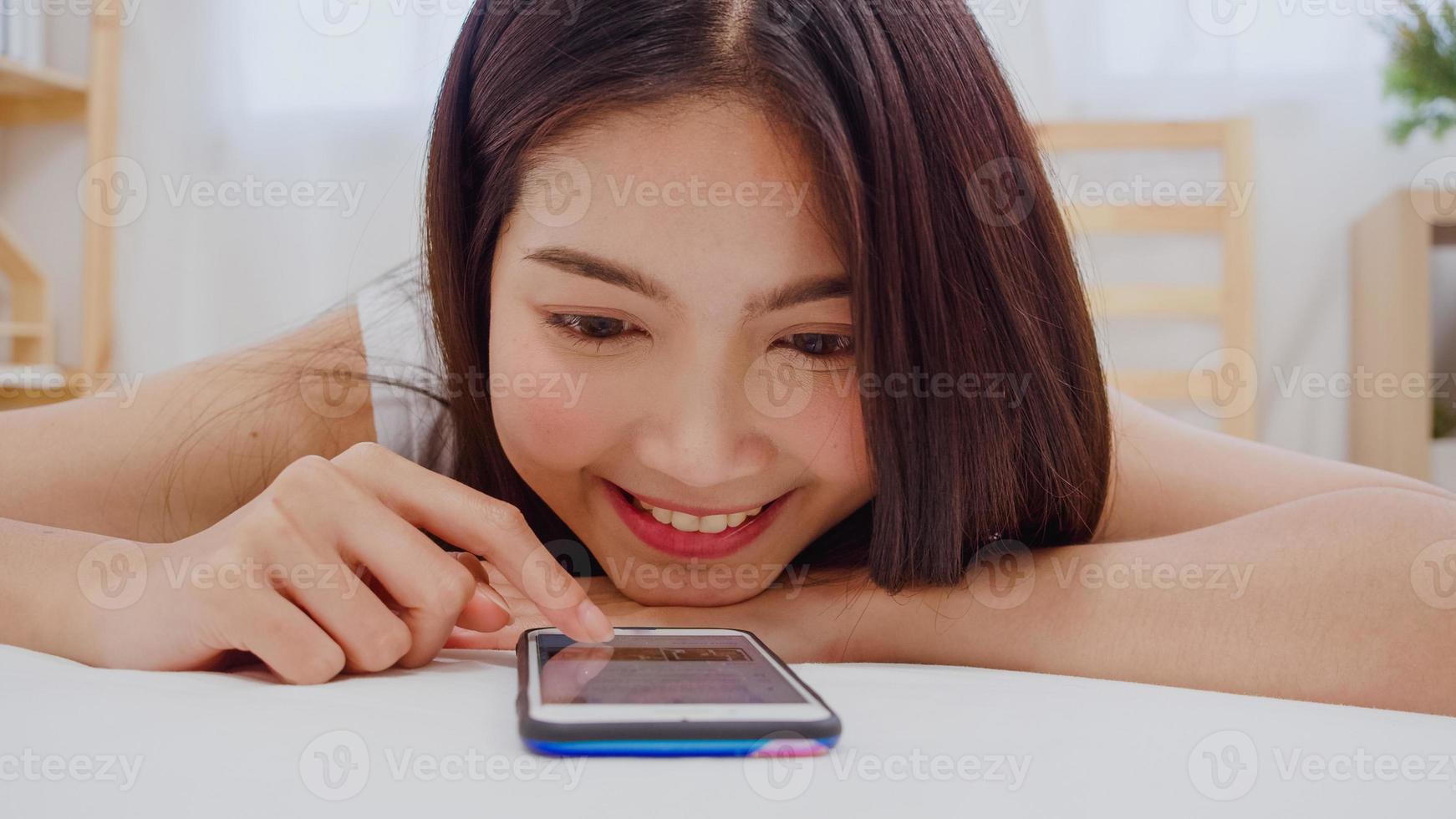 giovane donna asiatica che usa lo smartphone controllando i social media sentendosi felice sorridente mentre si trova sul letto dopo essersi svegliata al mattino, bella ragazza giapponese attraente sorridente si rilassa in camera da letto a casa. foto