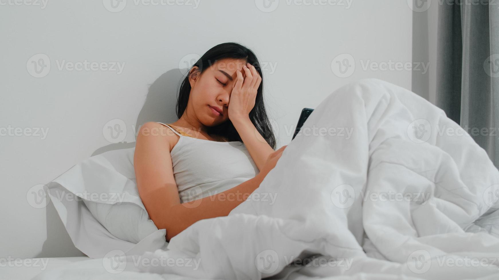 giovane donna asiatica che usa lo smartphone controllando i social media sentendosi felice sorridente mentre si trova sul letto dopo essersi svegliata al mattino, bella attraente signora ispanica sorridente si rilassa in camera da letto a casa. foto