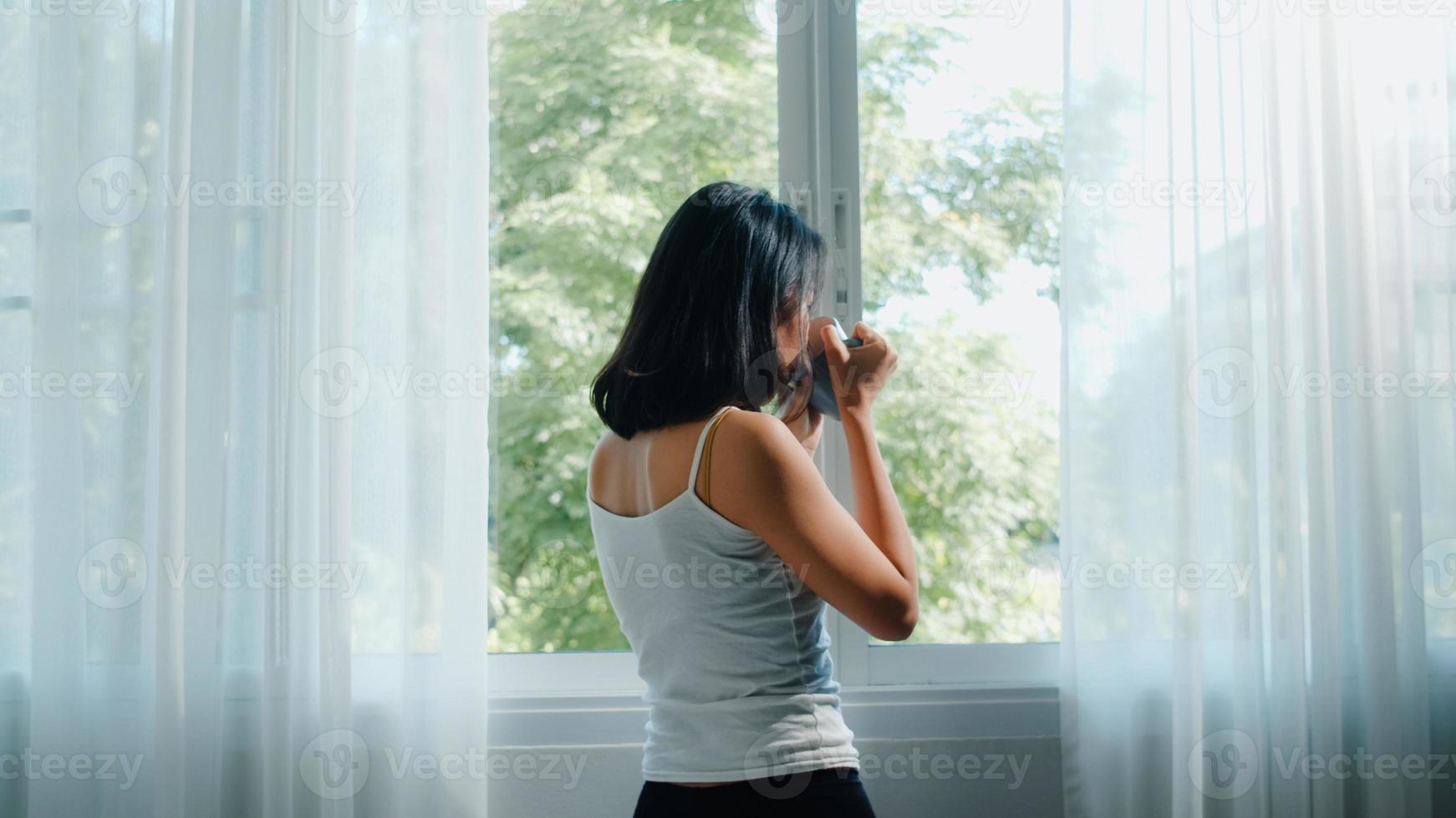 felice bella donna asiatica sorridente e bere una tazza di caffè o tè vicino alla finestra in camera da letto. la giovane ragazza latina apre le tende e si rilassa al mattino. stile di vita signora a casa concetto. foto