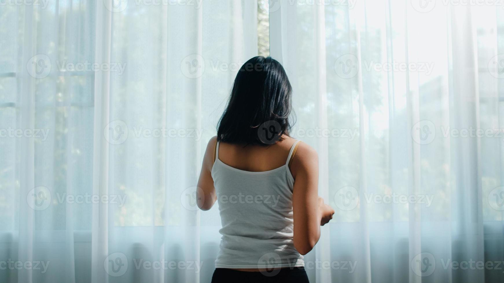 felice bella donna asiatica sorridente e bere una tazza di caffè o tè vicino alla finestra in camera da letto. la giovane ragazza latina apre le tende e si rilassa al mattino. stile di vita signora a casa concetto. foto