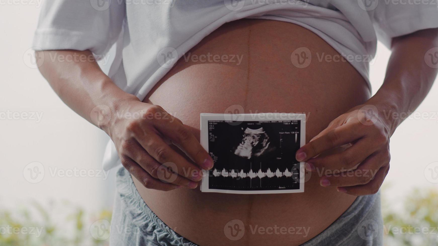 le giovani donne incinte asiatiche mostrano e guardano la foto degli ultrasuoni bambino nella pancia. mamma che si sente felice sorridente tranquillo mentre si prende cura del bambino sdraiato vicino alla finestra nel soggiorno a casa concetto.