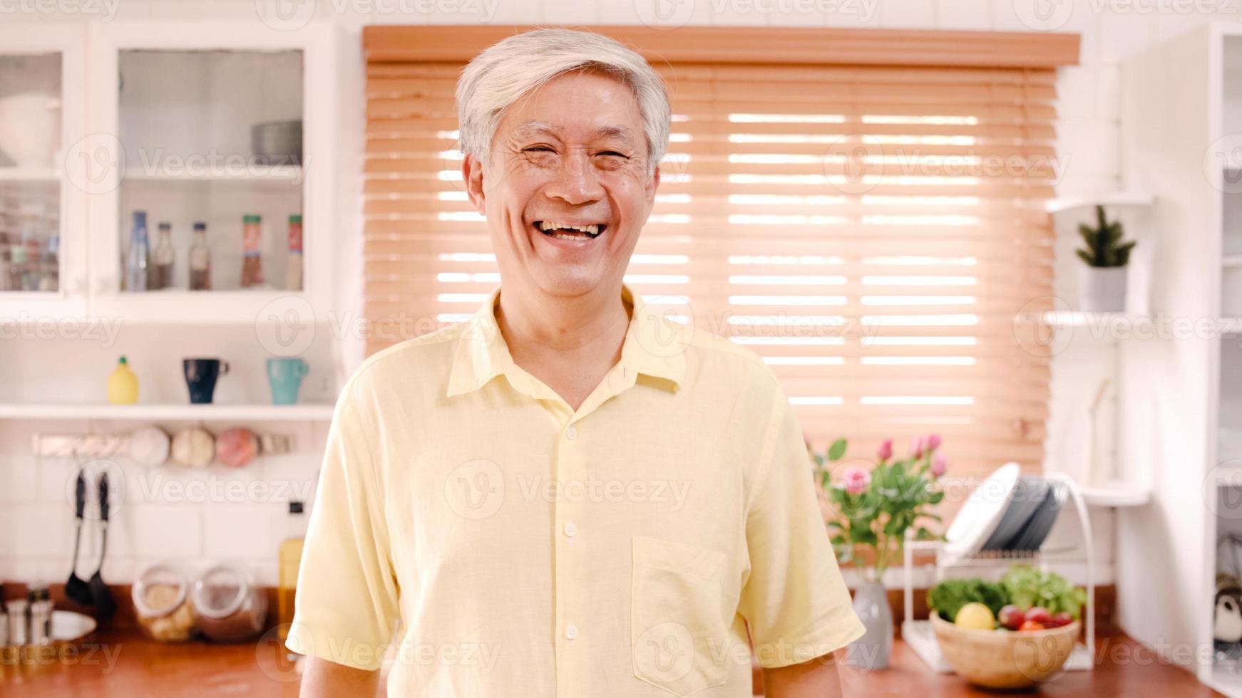 uomo anziano asiatico che si sente felice sorridendo e guardando la telecamera mentre si rilassa in cucina a casa. concetto di stile di vita degli uomini anziani a casa. foto