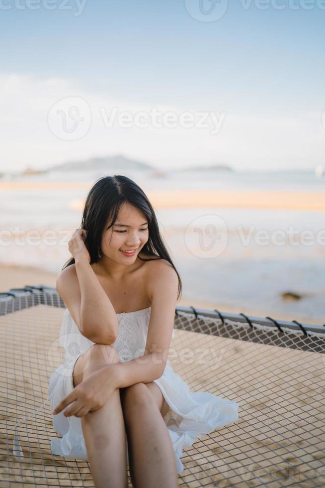 la giovane donna asiatica che si siede sull'amaca si rilassa sulla spiaggia, la bella femmina felice si rilassa vicino al mare. stile di vita le donne viaggiano sul concetto di spiaggia. foto