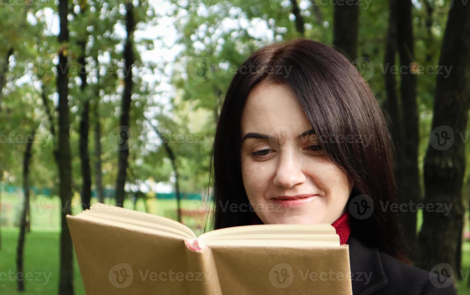 donna bruna sorridente che legge un libro nel parco. ritratto di una donna caucasica affascinante felice all'aperto. foto