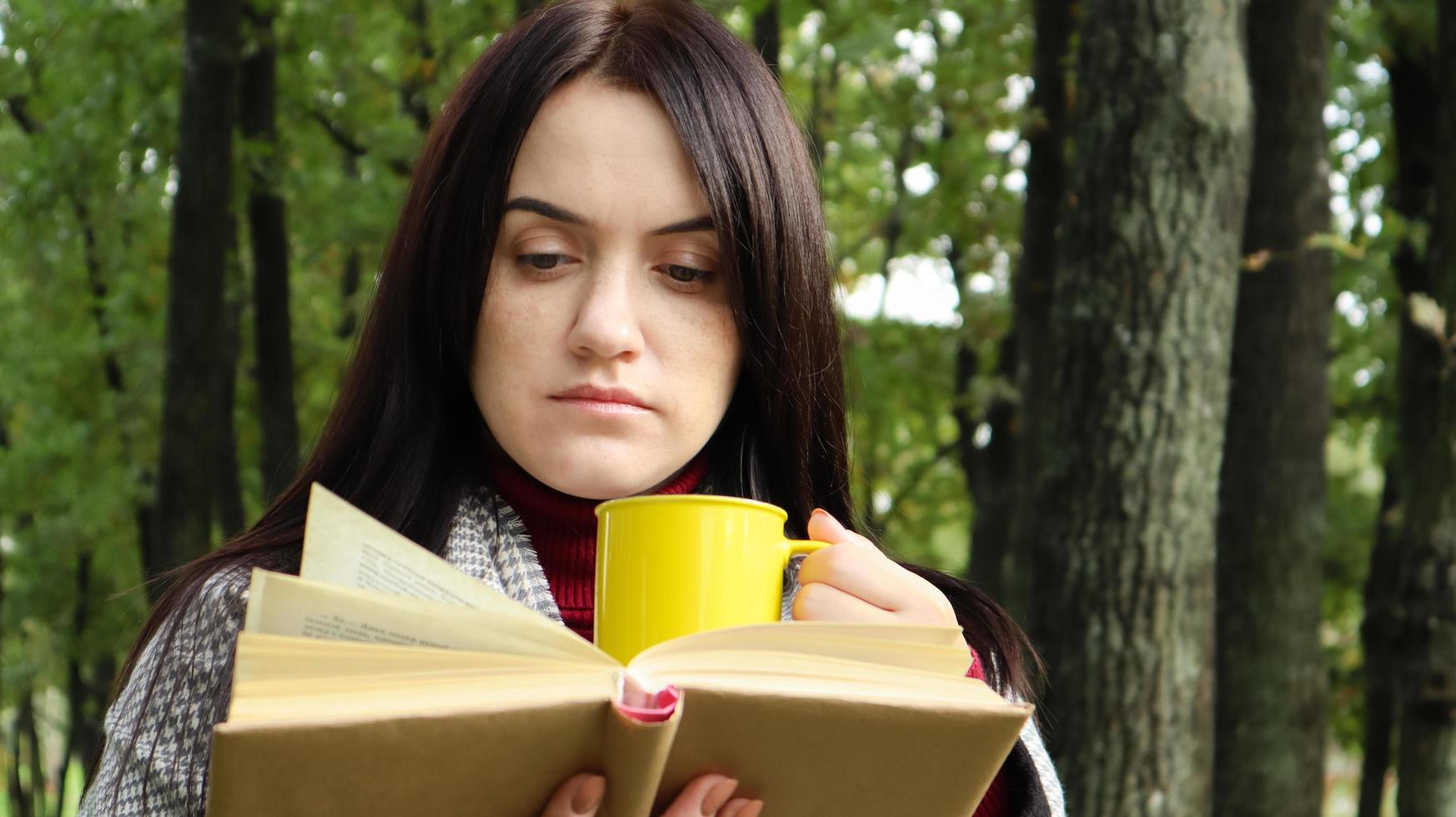 ritratto di una ragazza vestita con un cappotto e una sciarpa nella foresta autunnale legge un libro e tiene in mano una tazza con una bevanda calda in un parco cittadino in una calda giornata. concetto di lettura e relax. foto