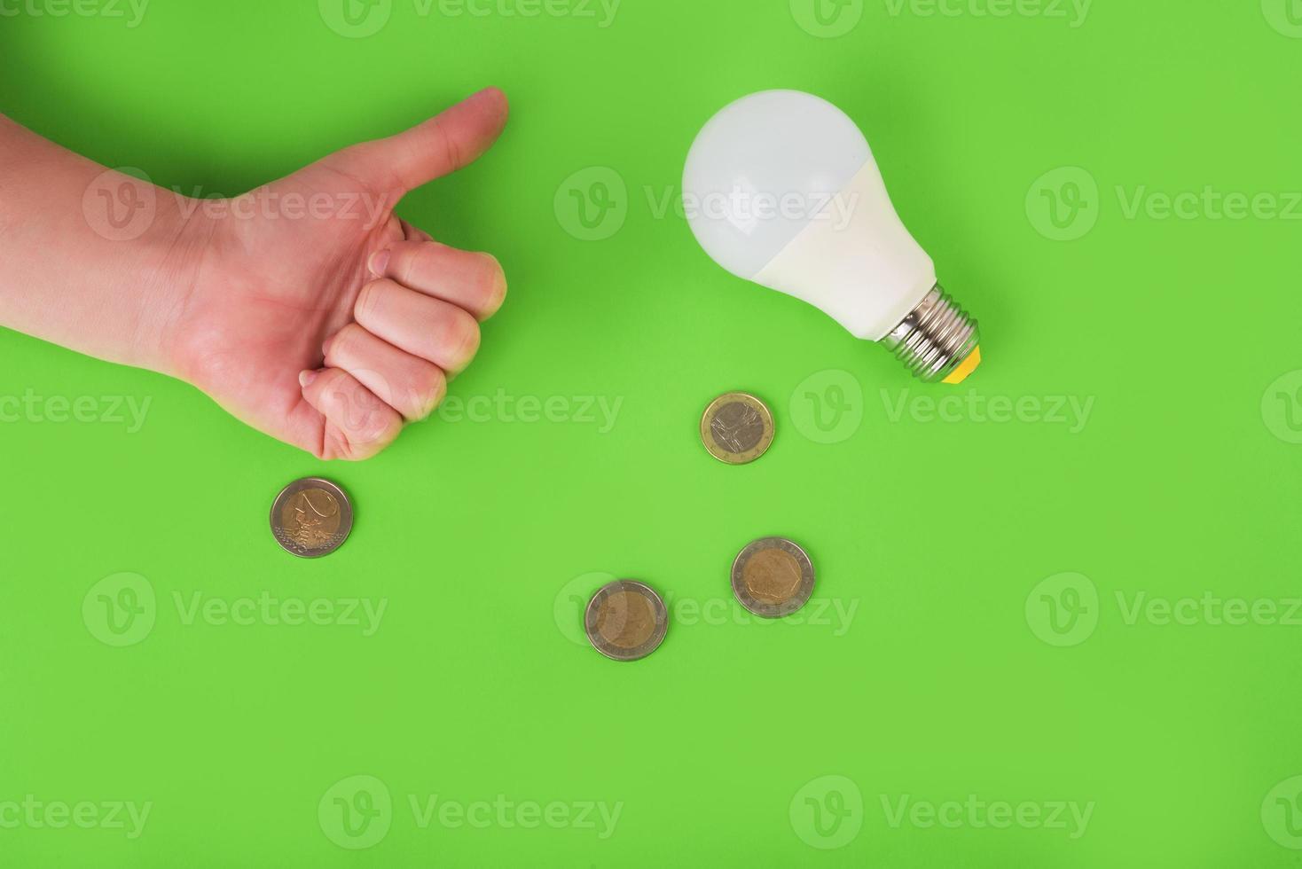 lampada a risparmio energetico, segno di approvazione a mano e monete in euro su sfondo verde. vista dall'alto. concetto di risparmio energetico. posto per il testo foto