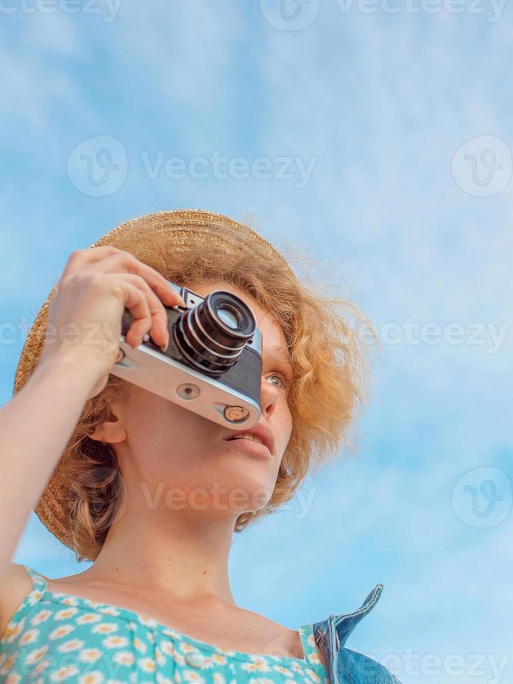 giovane donna rossa riccia con cappello di paglia, prendisole blu e giacca di jeans in piedi con una fotocamera vintage e scattare foto su sfondo blu cielo. divertimento, estate, moda, riprese, viaggi, concetto di gioventù