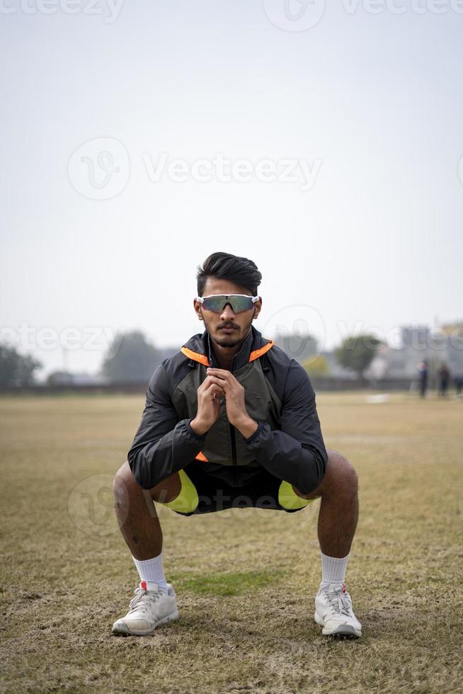 colpo frontale di un ragazzo indiano che fa squat nel campo indossando abbigliamento sportivo. concetto di sport e stile di vita. foto