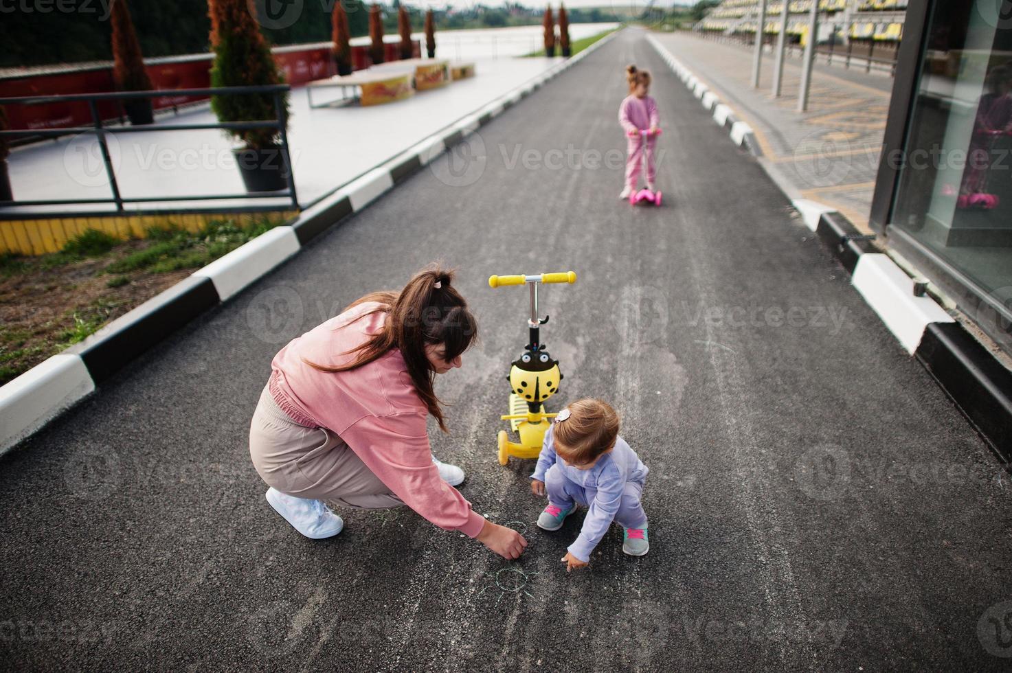 giovane madre alla moda con due ragazze all'aperto. la famiglia sportiva trascorre il tempo libero all'aria aperta con gli scooter. dipinto con il gesso sull'asfalto. foto