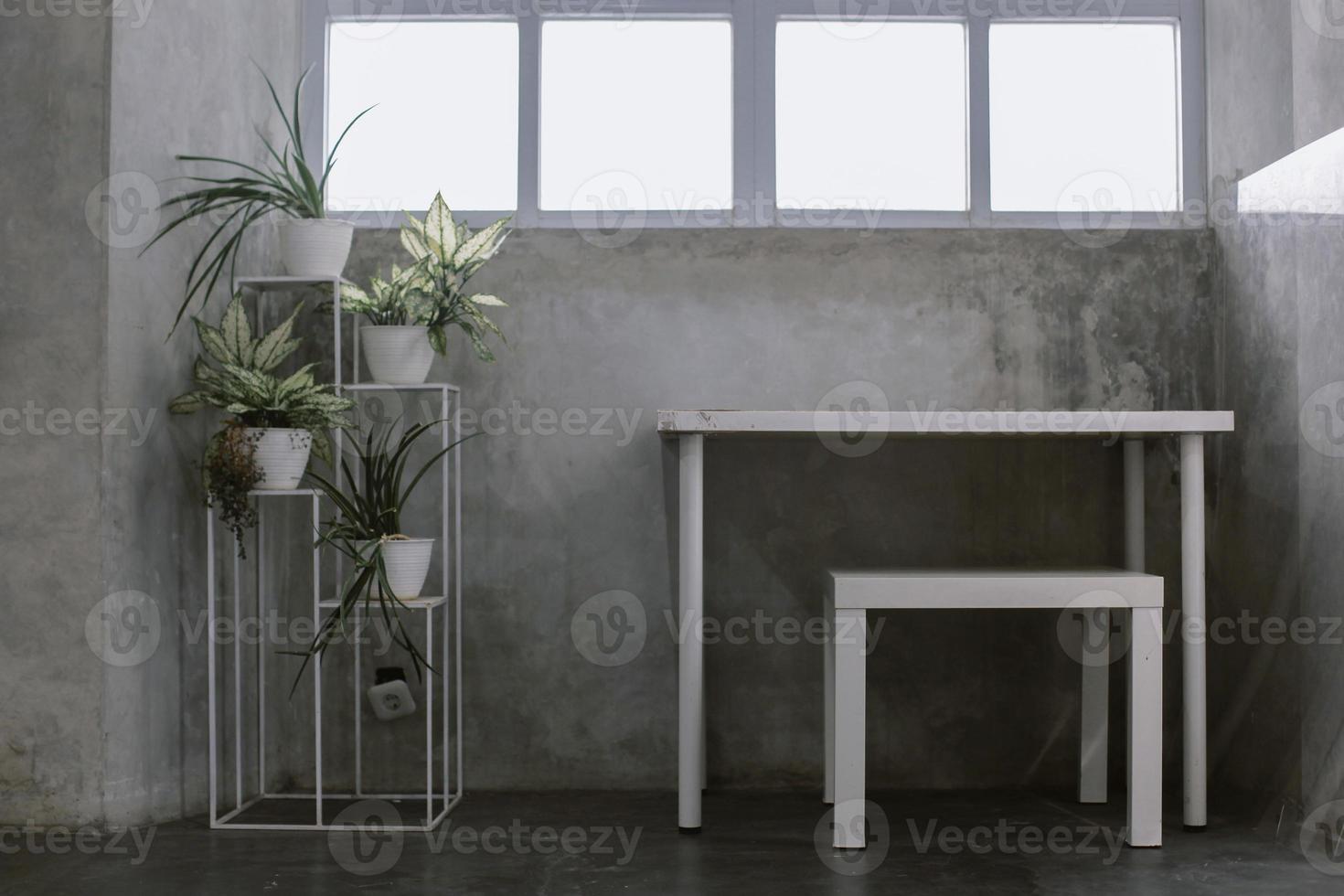 concetto di interni industriali minimalisti con set da scrivania in legno bianco, rack bianco di piante in vaso e pareti in cemento grunge con finestre quadrate bianche per l'illuminazione foto