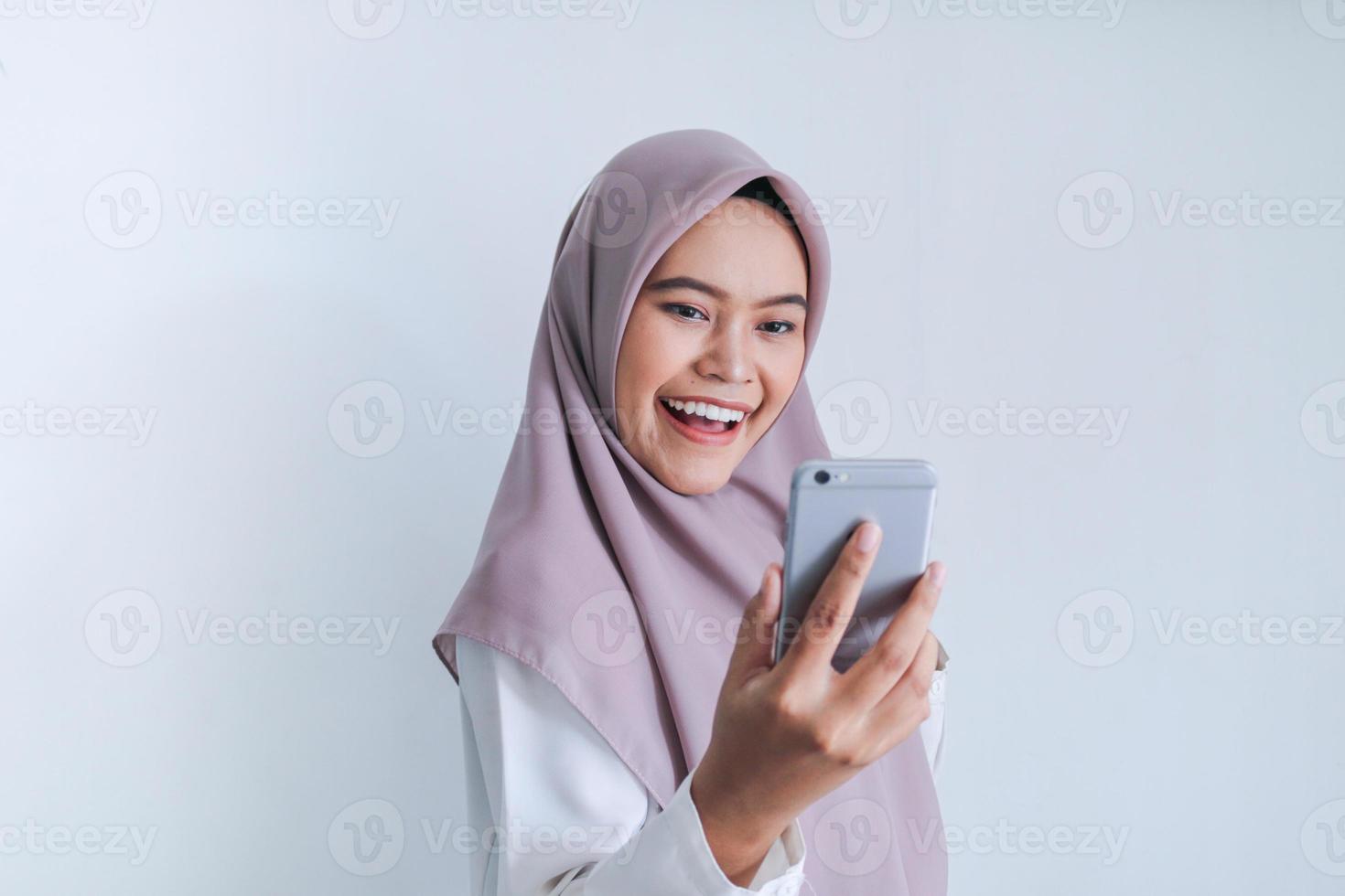 la giovane donna asiatica dell'islam che indossa il velo è felice ed entusiasta di celebrare in ciò che vede sullo smartphone. donna indonesiana su sfondo grigio foto